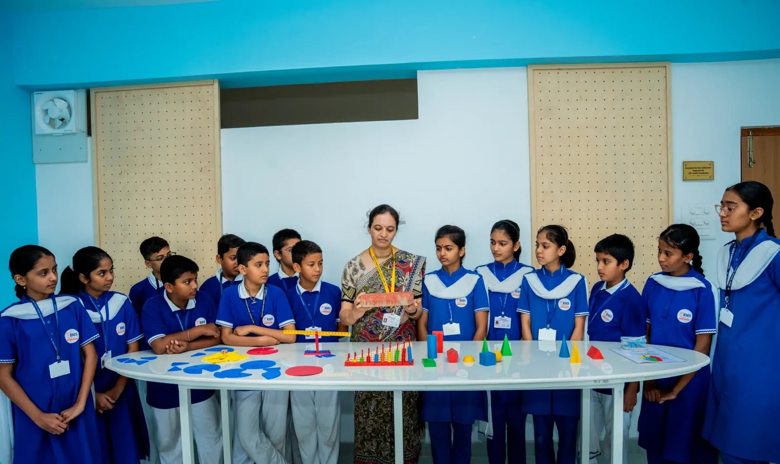 A class around the activity table with their teacher and shape and number material