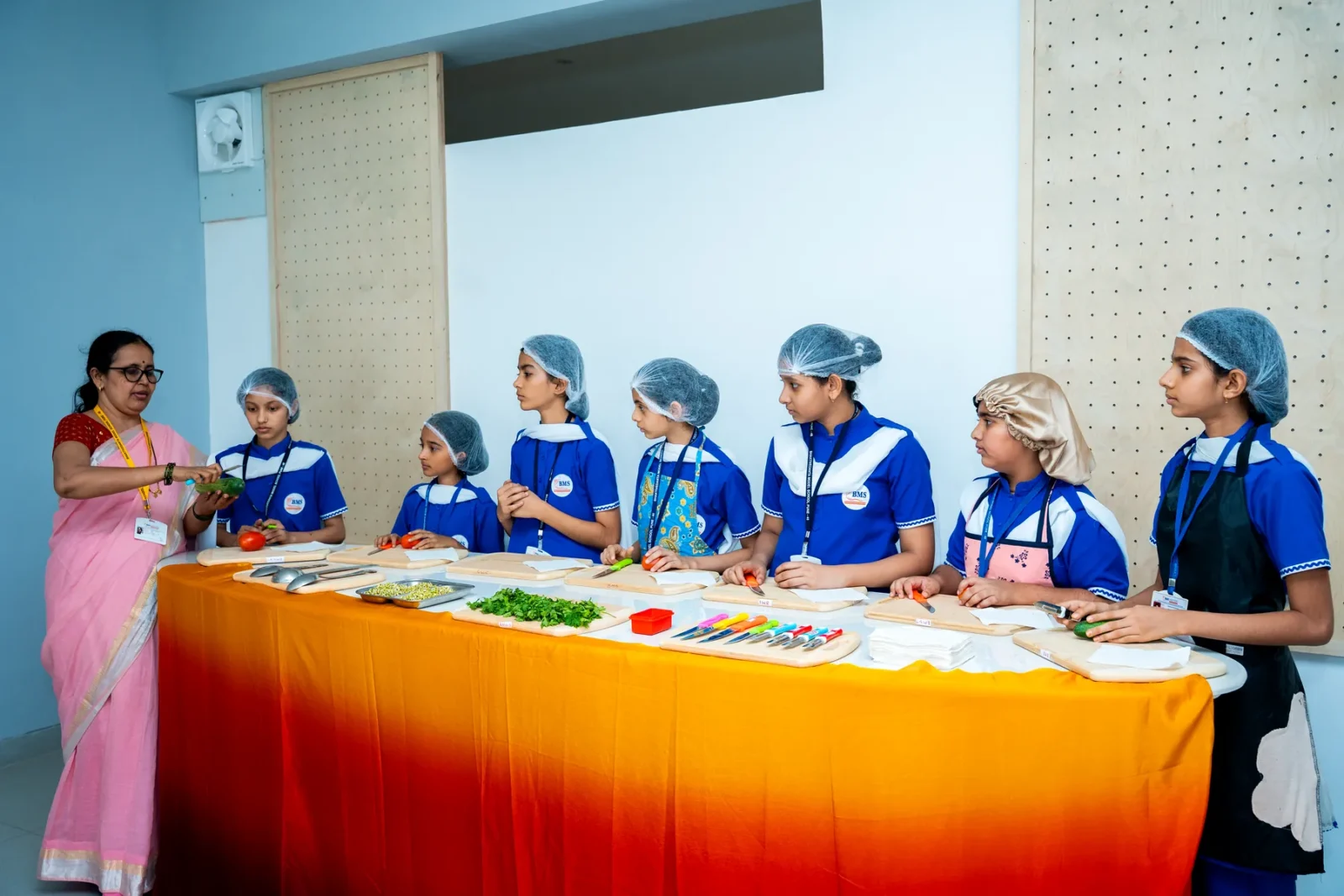 Students in caps and aprons at a fireless cooking class, preparing vegetables at a long table with their teacher