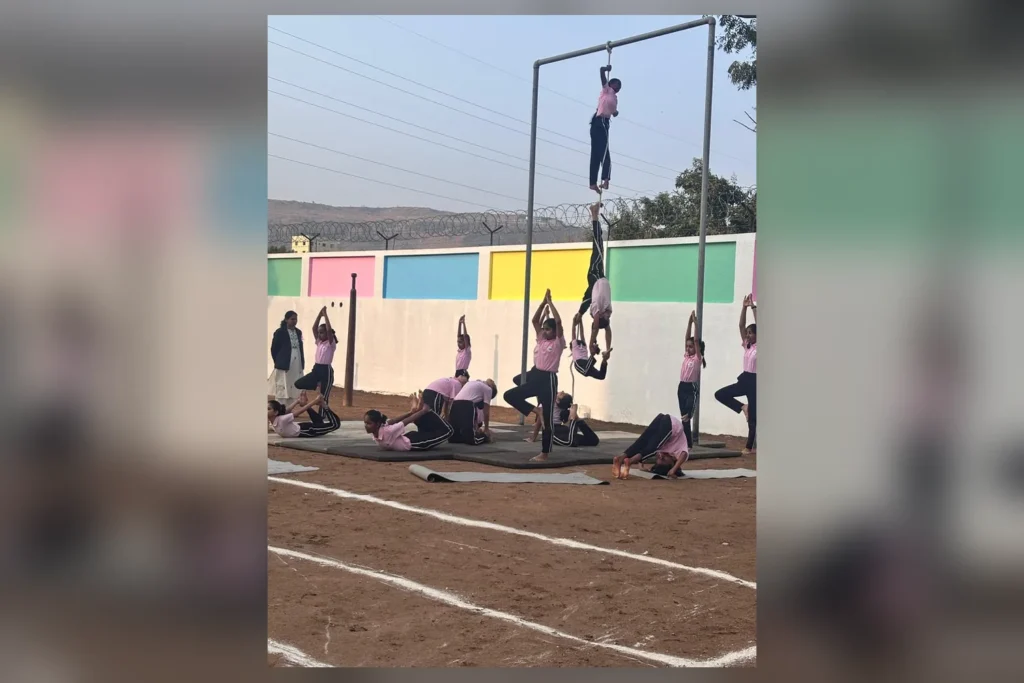A student climbing the mallakhamb pole while the class practises on mats below
