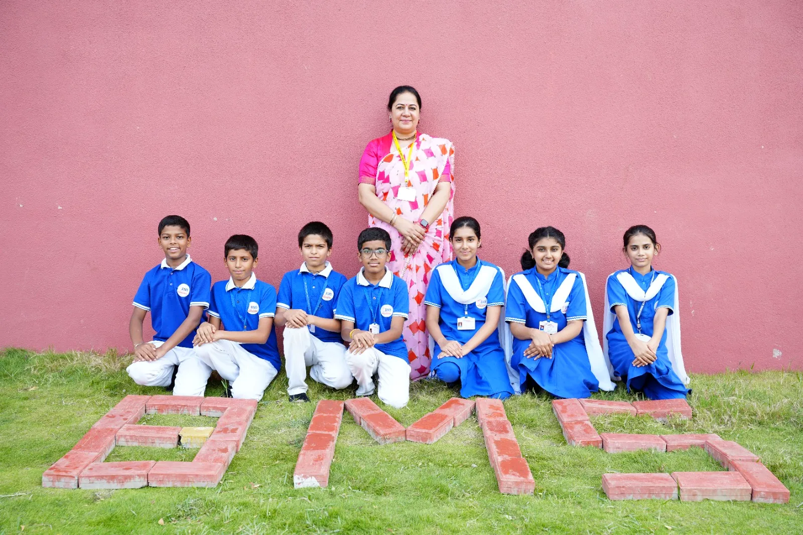 Students in school uniform with their class teacher, the letters B M S laid out in brick on the grass in front of them