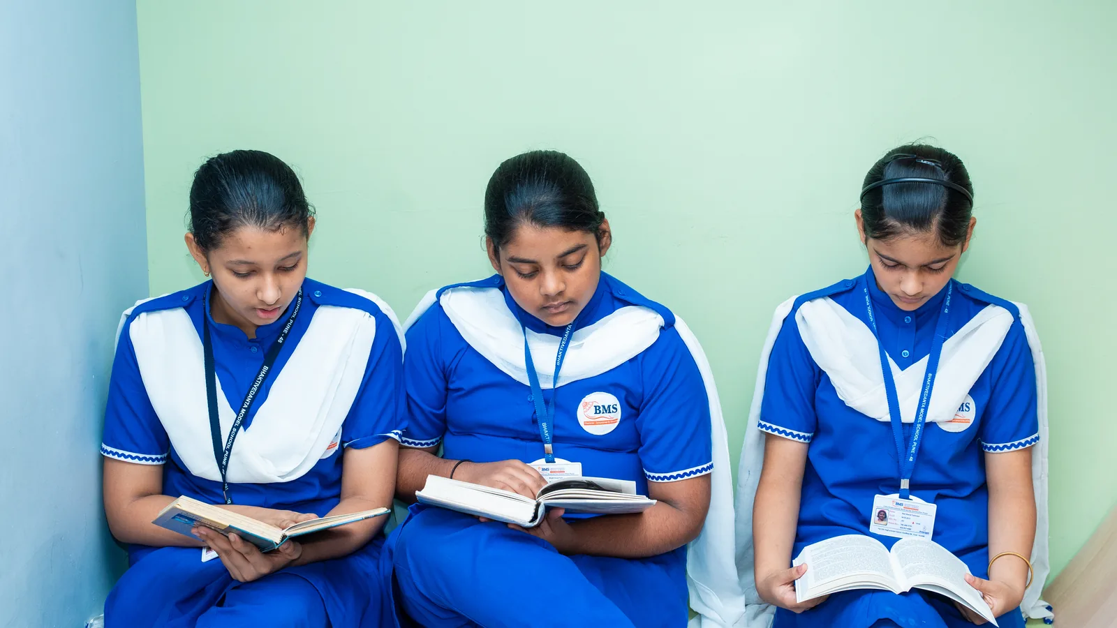 Three students reading in school uniform, seated against a green wall